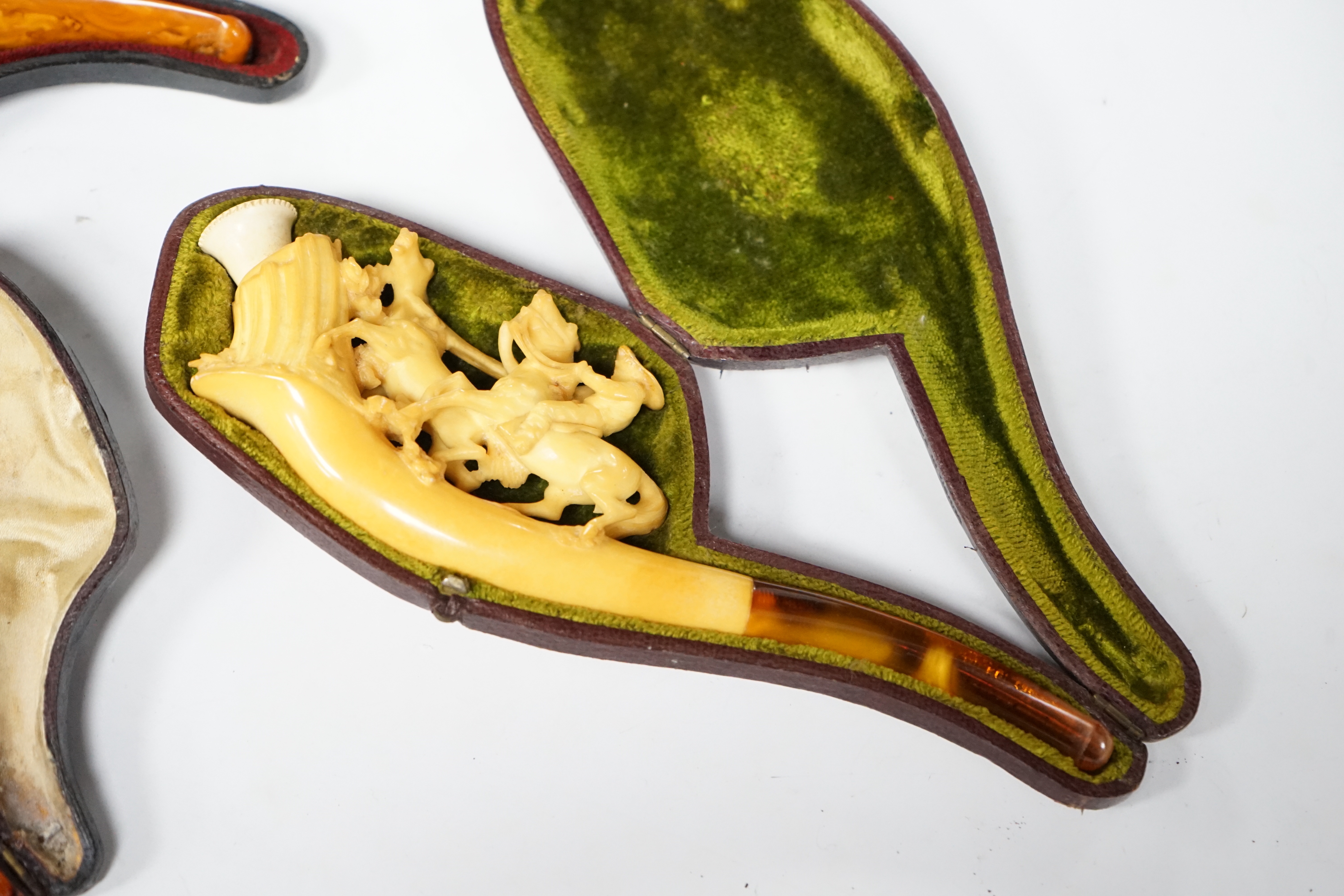 Three cased novelty Meerschaum pipes and pipe bowls, all with amber mouth pieces, one of a horses and cowboy, another a model of a hand and the other of a scantily clad lady wearing a riding cap, largest 17 cm long